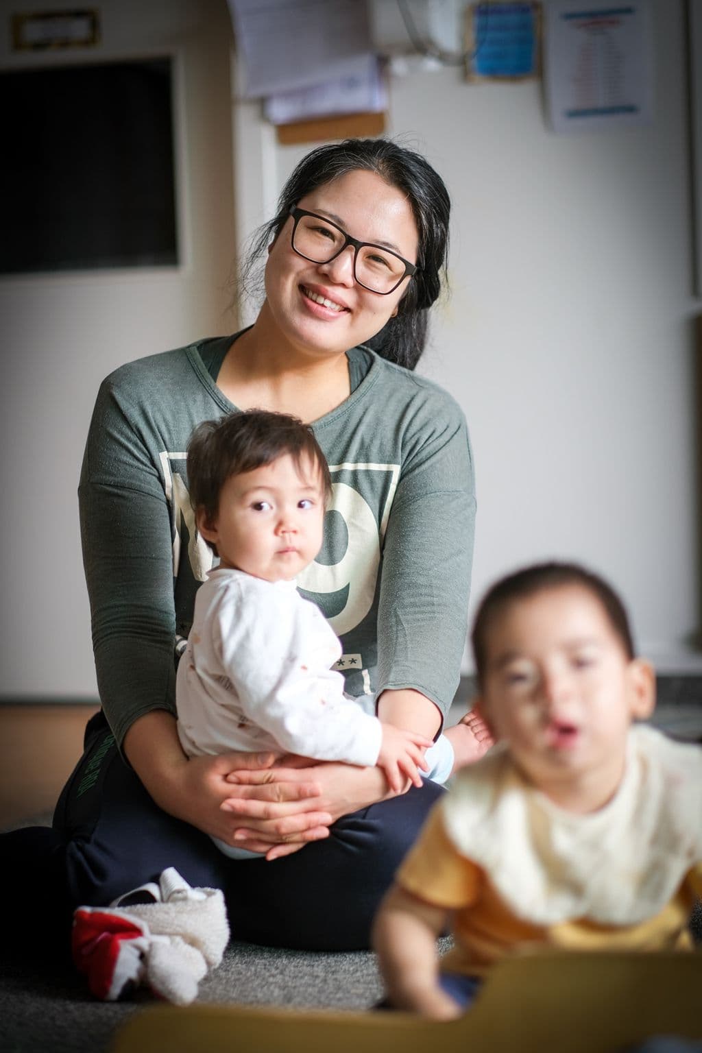 Infant kaiako holding a pēpi on her lap while supervising another toddler in the nursery at Triton Montessori Educare